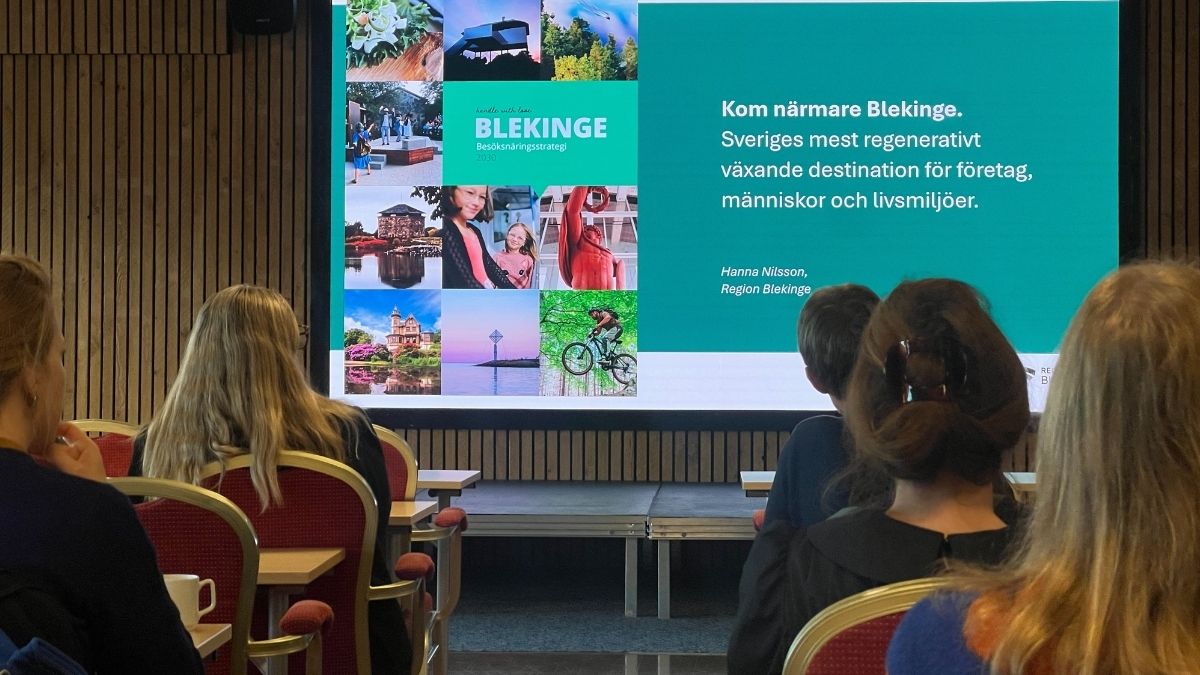 Deltagare lyssnar på presentation om Blekinges besöksnäringsstrategi med budskapet "Kom närmare Blekinge" på storbildsskärm.