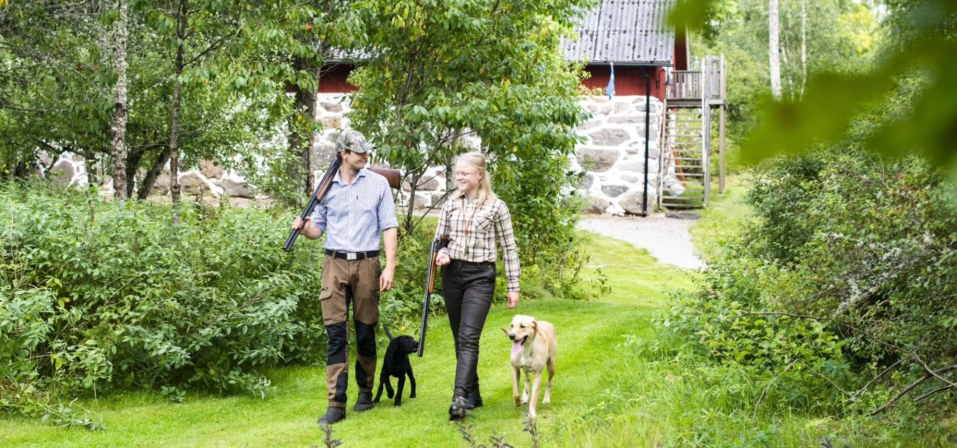 Två jägare med gevär och hundar promenerar genom grönskande natur vid en röd jaktstuga i Olofström, Blekinge.
