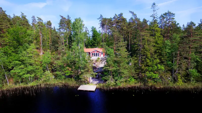 Röd stuga vid sjö i skogsmiljö i Olofström, med brygga och omgiven av tät grönska.
